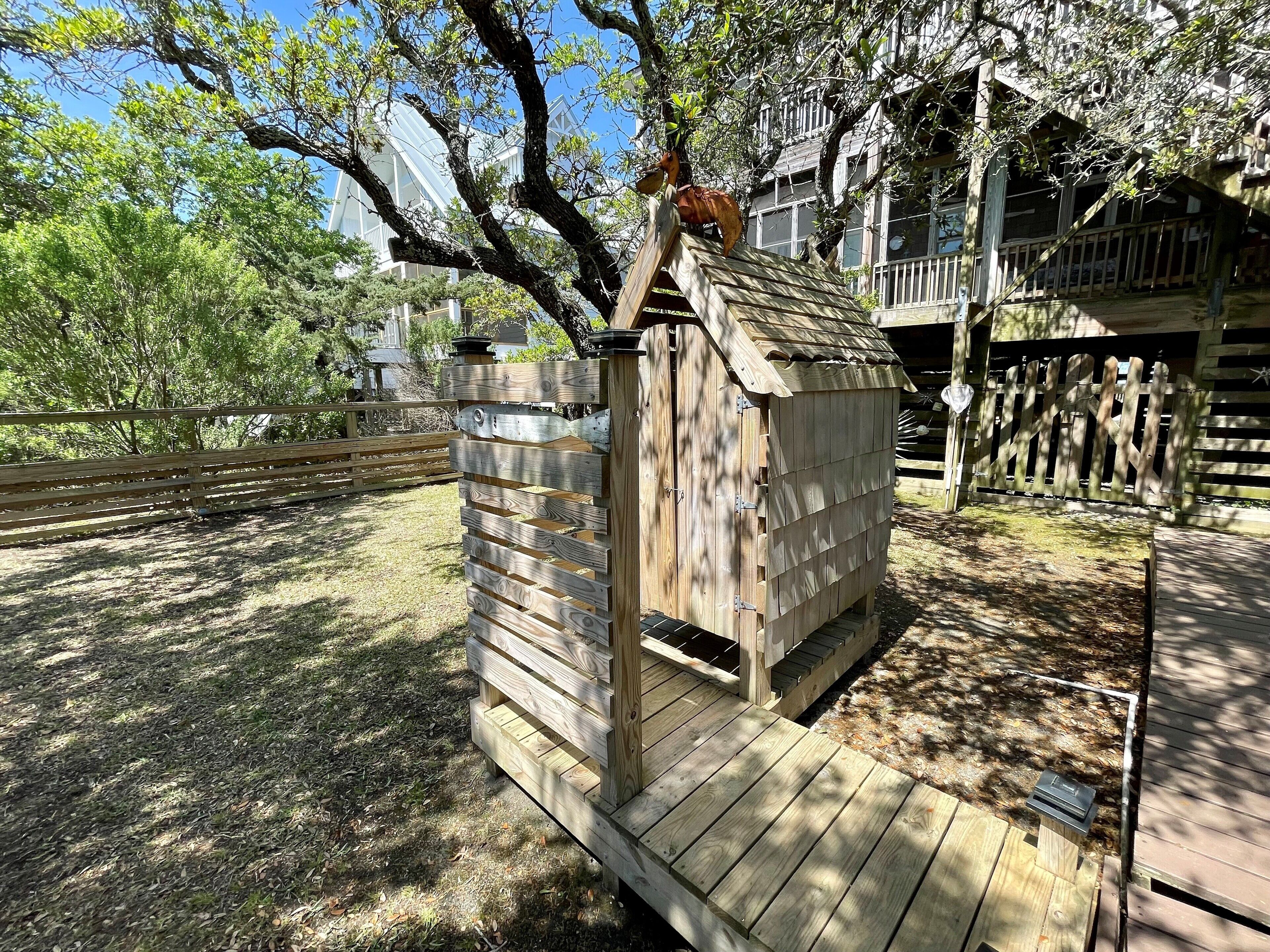A wooden structure with a sloped roof, possibly a shed or a small building, situated in a fenced outdoor area with trees and a building in the background.