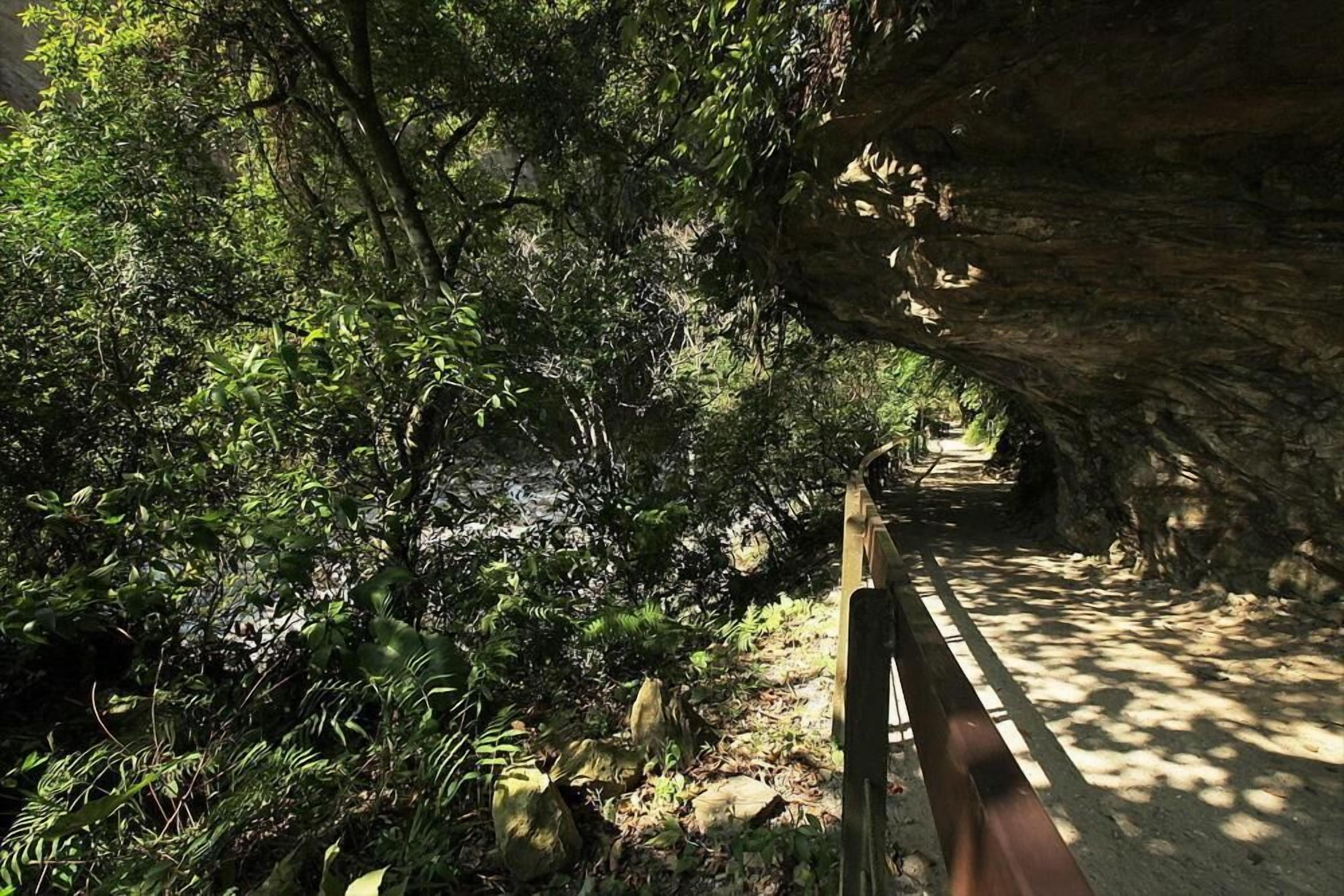 A pathway through a forested area with a wooden railing on one side and a rocky overhang on the other.