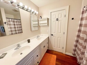 A bathroom with a double sink, a large mirror with lights, a door with a handle, and a shower curtain. - Redbeard's Claim: Professionally decorated, perfect relaxing island home. (Ocracoke)