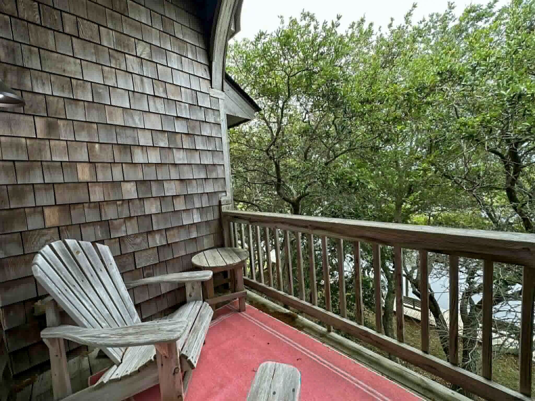 A wooden deck with Adirondack chairs, a small table, and a railing overlooking a body of water.