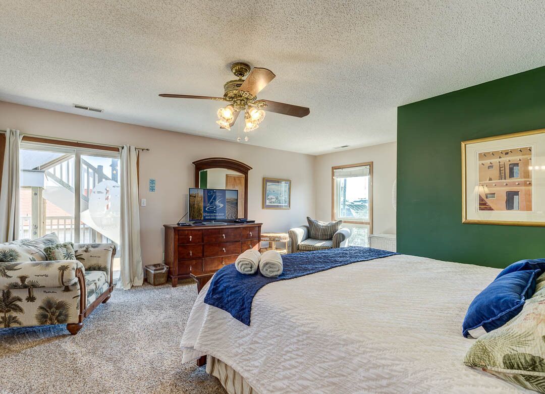 A bedroom with a large bed, a ceiling fan, a dresser, a chair, and a view of the outside.