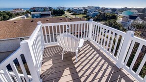 A wooden deck with a white railing, a white plastic chair, and a view of houses and the ocean.