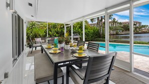 A covered outdoor dining area with a pool view, a grill, and lush greenery.