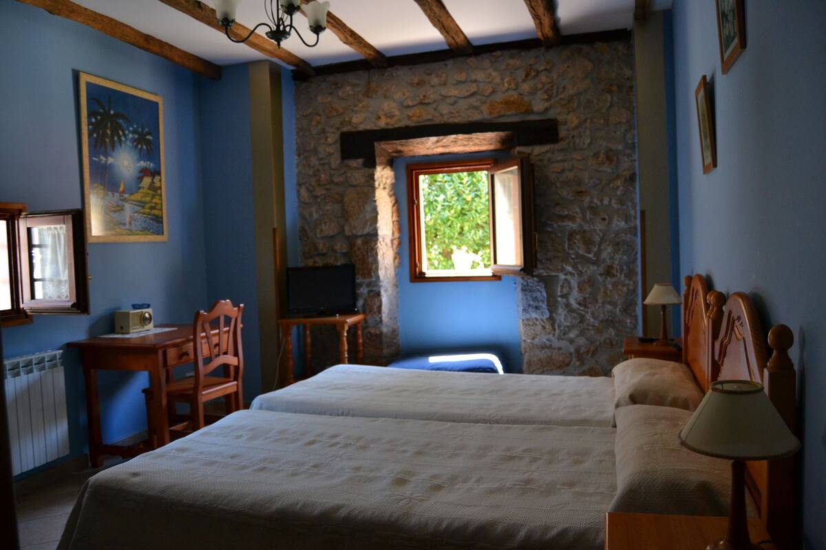 Quarto com duas camas, uma escrivaninha de madeira, televisão e parede de pedra.