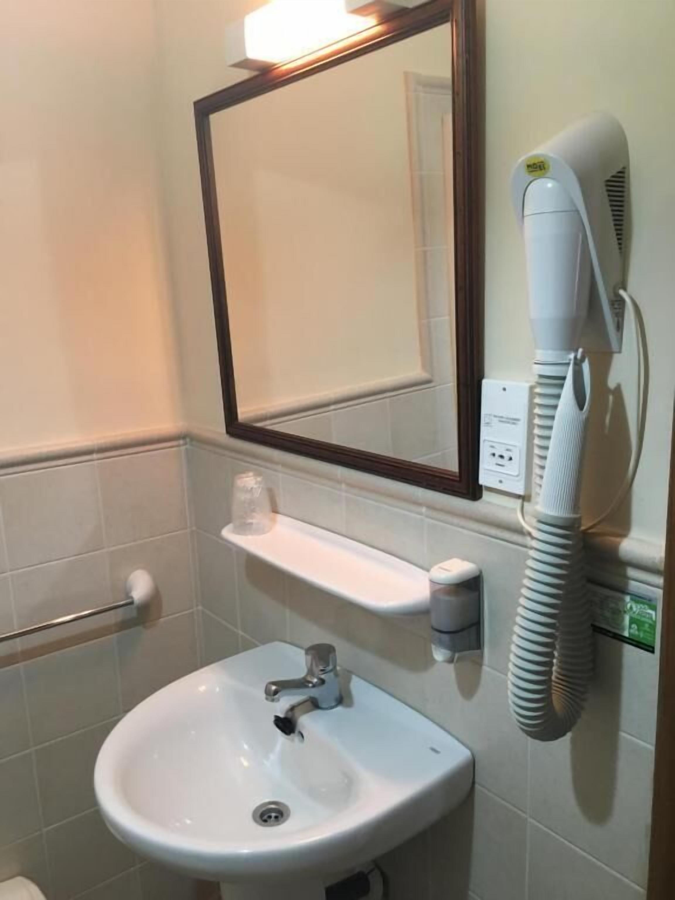 Une salle de bain avec un lavabo blanc, un miroir, un porte-serviettes, un sèche-cheveux et une petite étagère sur laquelle se trouvent un verre et un récipient.