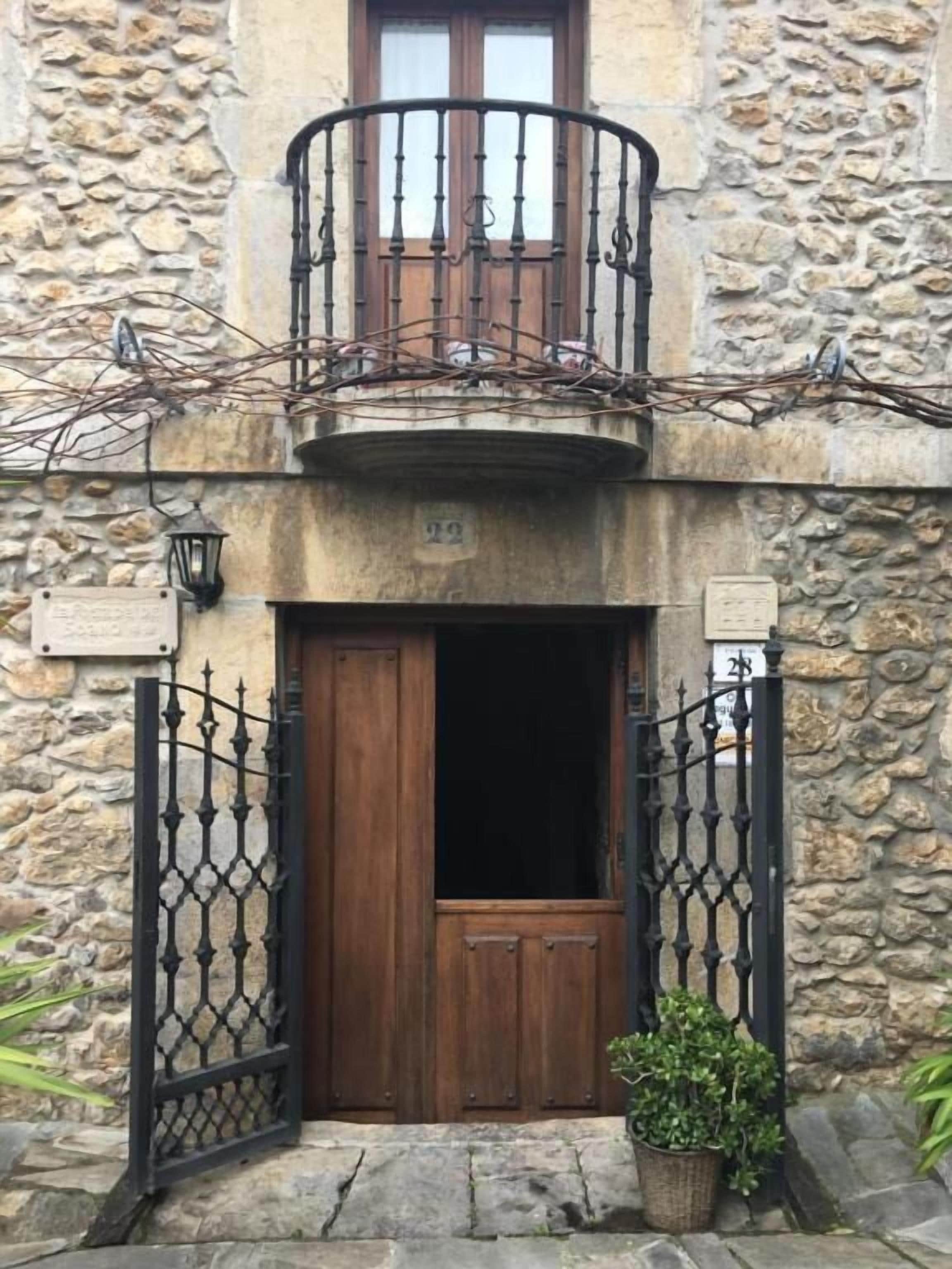 A stone building with a wooden door, a balcony with wrought iron railings, and a potted plant in front.