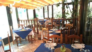 A restaurant with multiple tables and chairs, covered by a wooden canopy, and surrounded by greenery. - Posada Soano (Arnuero)