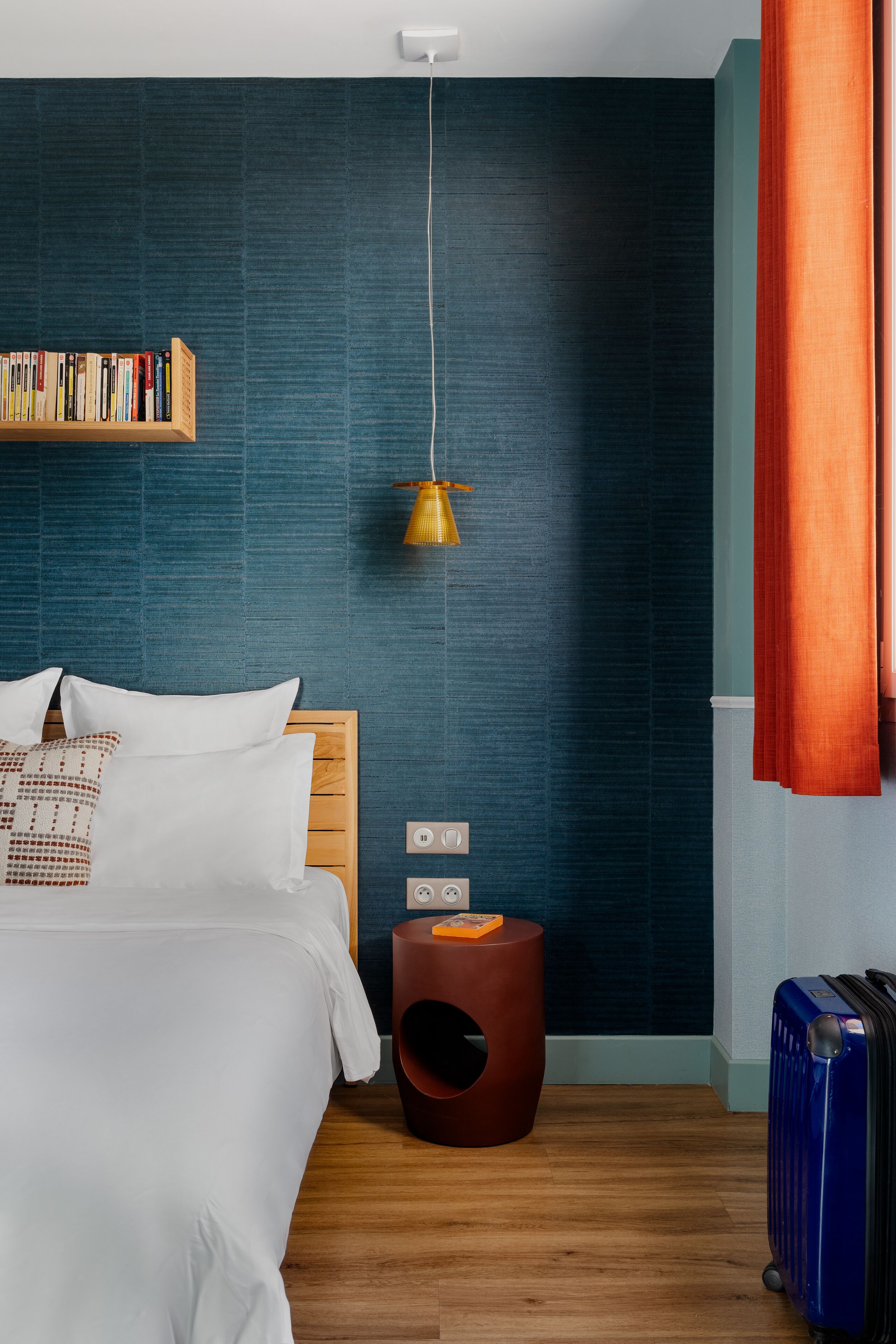 Une chambre d’hôtel moderne avec un lit en bois, une bibliothèque, un tabouret rond original et une valise.