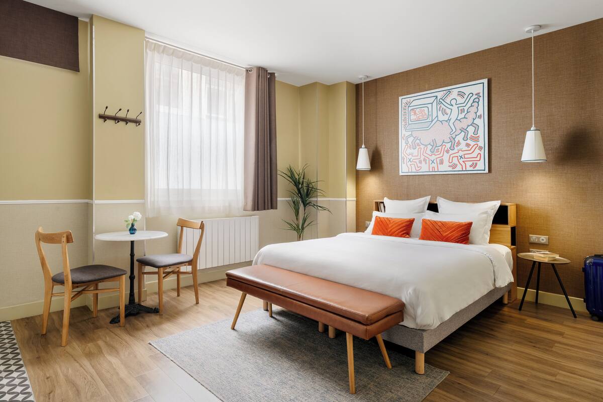 Una habitación de hotel moderna con una cama grande, un banco, dos sillas, una mesita y una planta en maceta.