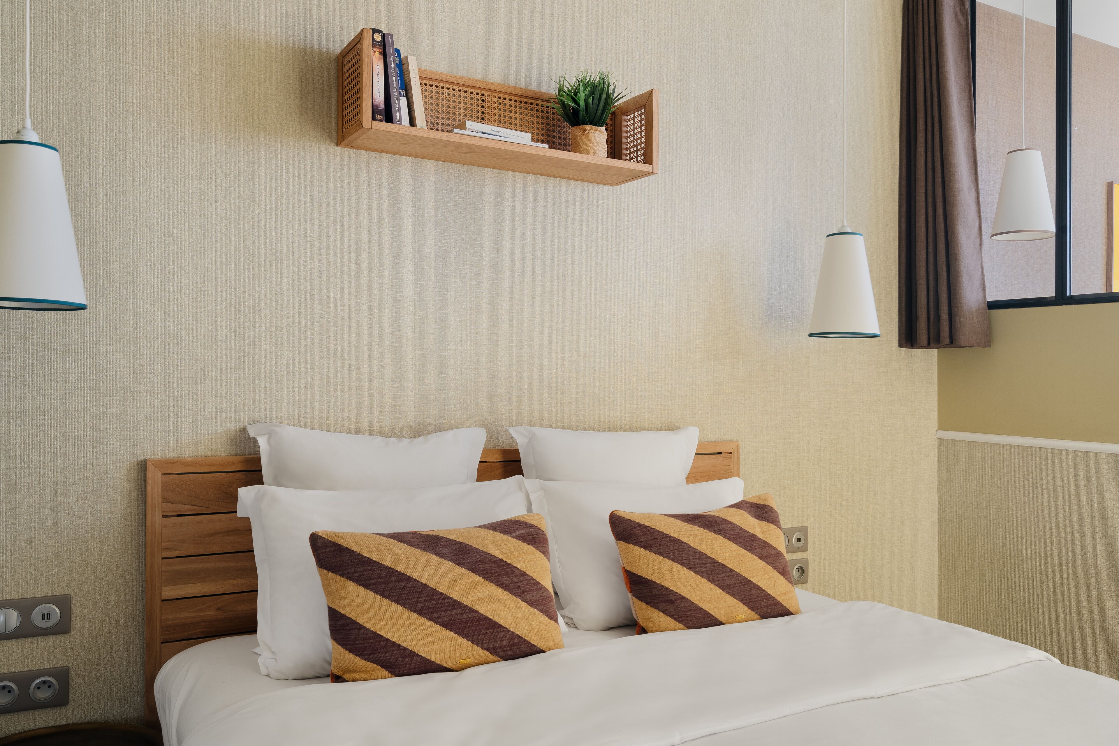 A neatly made bed with white and striped pillows, a wooden headboard, and a shelf above holding books and a plant.