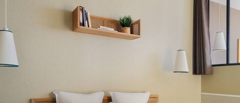 A neatly made bed with white and striped pillows, a wooden headboard, and a shelf above holding books and a plant.