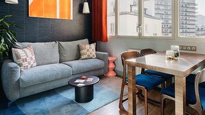 Een moderne woonkamer met een grijze bank, een houten eettafel met blauwe stoelen, een klein roze bijzettafeltje en een groot raam met gordijnen.