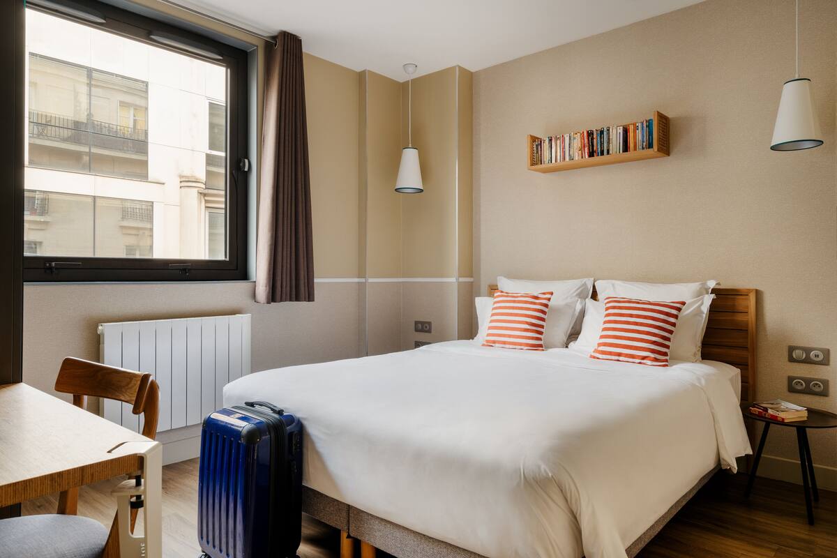 Habitación de hotel con una cama grande, cabecero de madera, mesita de noche, una silla, radiador, ventana con cortinas y una estantería con libros.