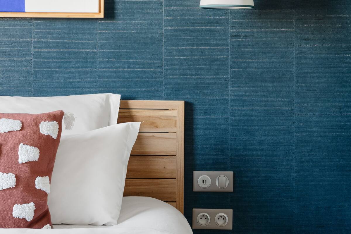 Habitación de hotel ordenada con cama, almohadas, una obra de arte enmarcada, una lámpara colgante, una mesita con un libro y enchufes en la pared.