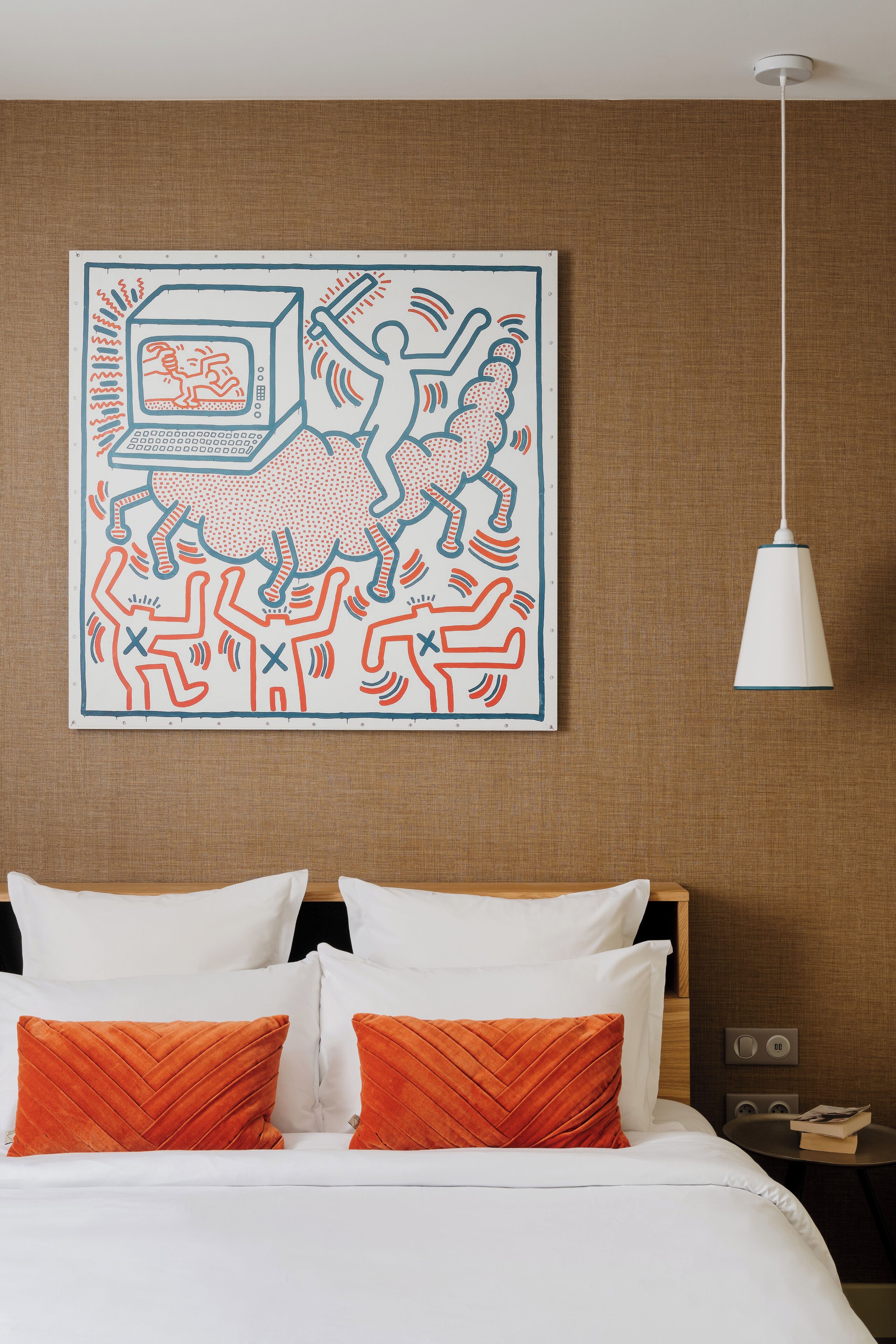 Ett hotellrum med en säng, två orange kuddar, en vit lampa och ett konstverk som hänger på väggen.