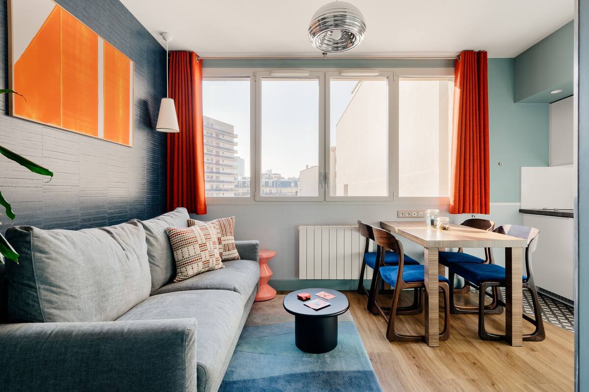 Una sala de estar moderna con un sofá gris, una mesa de centro redonda y un comedor con una mesa de madera y sillas azules.