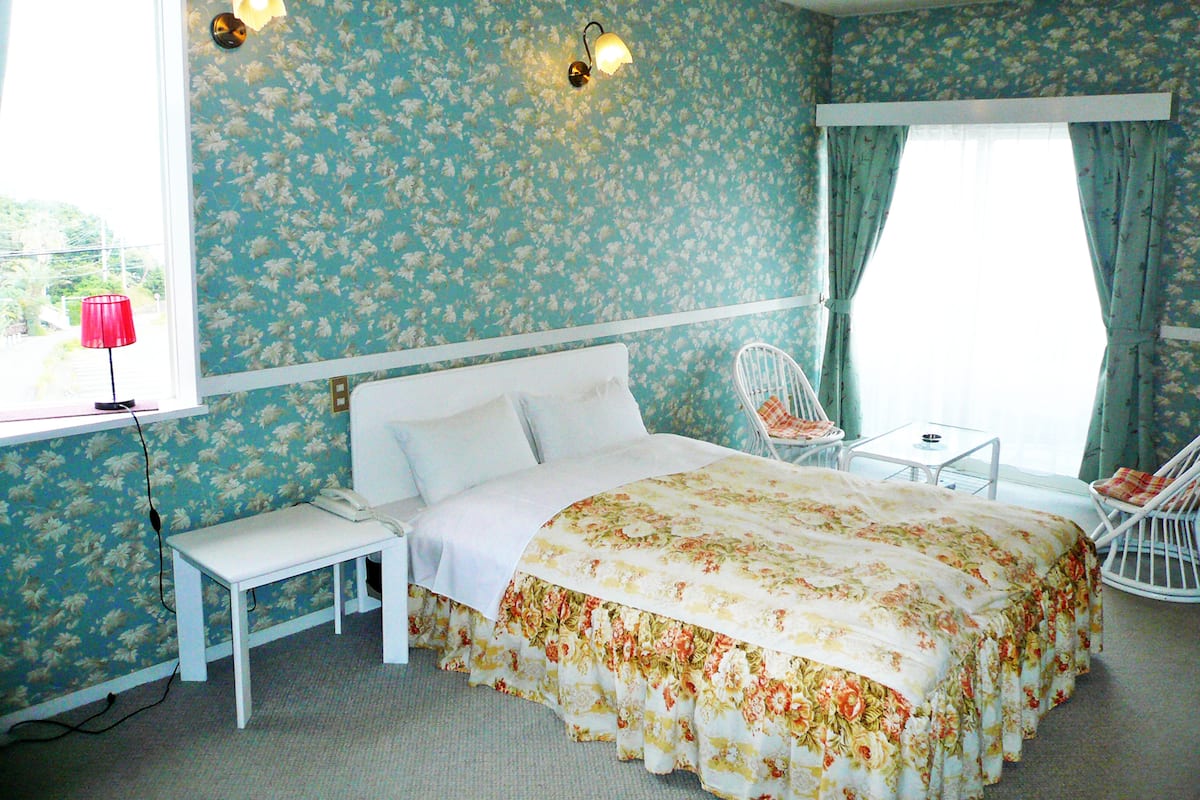A bedroom with floral wallpaper, a bed with a floral bedspread, a bedside table, a chair, and a window with curtains.