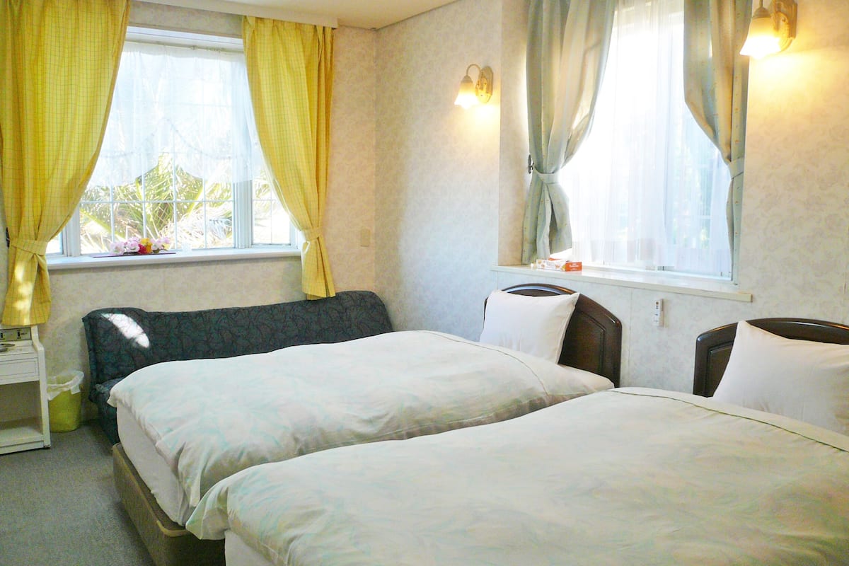 A hotel room with two beds, a nightstand, a lamp, and a window with curtains.