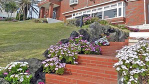 A two-story brick house with a front porch, a small garden with pink and white flowers, and a palm tree in the background. - Pension A Million Roses (Shimoda)