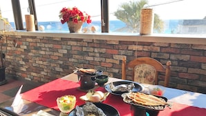 A table set outdoors with a brick wall backdrop, a wicker basket with flowers, and a view of the sea through large windows. - Pension A Million Roses (Shimoda)