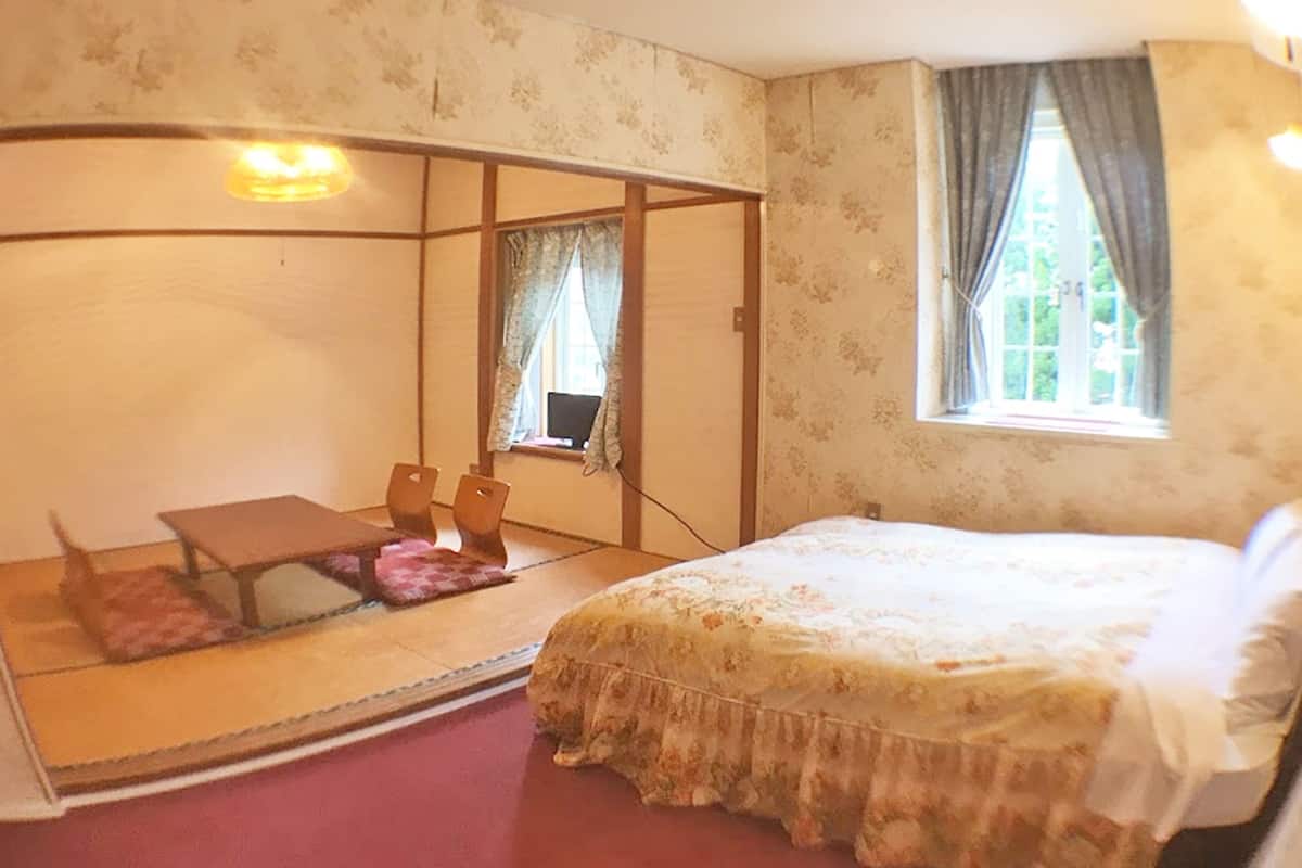 A bedroom with a bed, a desk, and a chair. There is a window with curtains and a view of greenery outside.