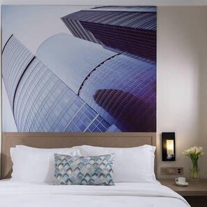 Habitación de hotel con una amplia ventana que ofrece vistas a edificios modernos, una cama bien hecha con sábanas blancas y una mesita de noche con una lámpara y un florero.