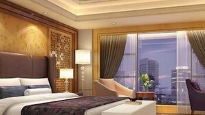 A hotel room with a large bed, a sitting area with a chair, a desk with a lamp, and a view of the city through large windows.