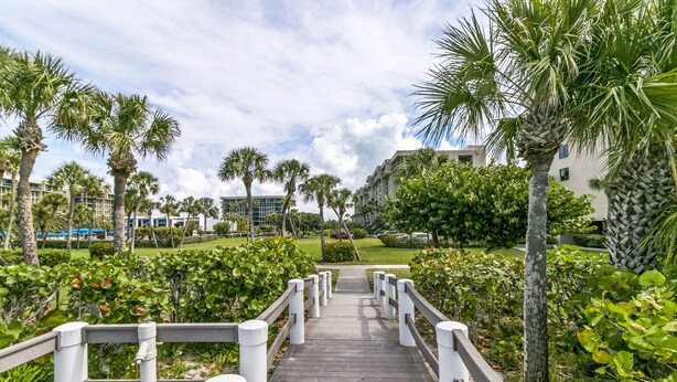 A wooden pathway leading to a building with a white railing, surrounded by palm trees and greenery. - Gulf-Front Luxury: 2BR Beachplace Condo on Longboat Key Unit 9-103 by RVA (Longboat Key)