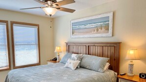 Ein Schlafzimmer mit einem Bett, zwei Nachttischen mit Lampen, einem Deckenventilator und einem Wandbild mit Strandmotiv.