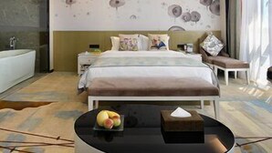 Une chambre d’hôtel moderne avec un grand lit, une table de chevet, une petite table avec des fruits, une œuvre d’art accrochée au mur et une grande fenêtre avec des rideaux.