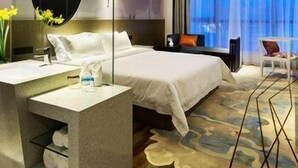 Une chambre d’hôtel avec un grand lit, une table de chevet avec un miroir, un petit bureau et une chaise. Il y a une fenêtre avec des rideaux, un tapis et un mur orné d’un tableau.