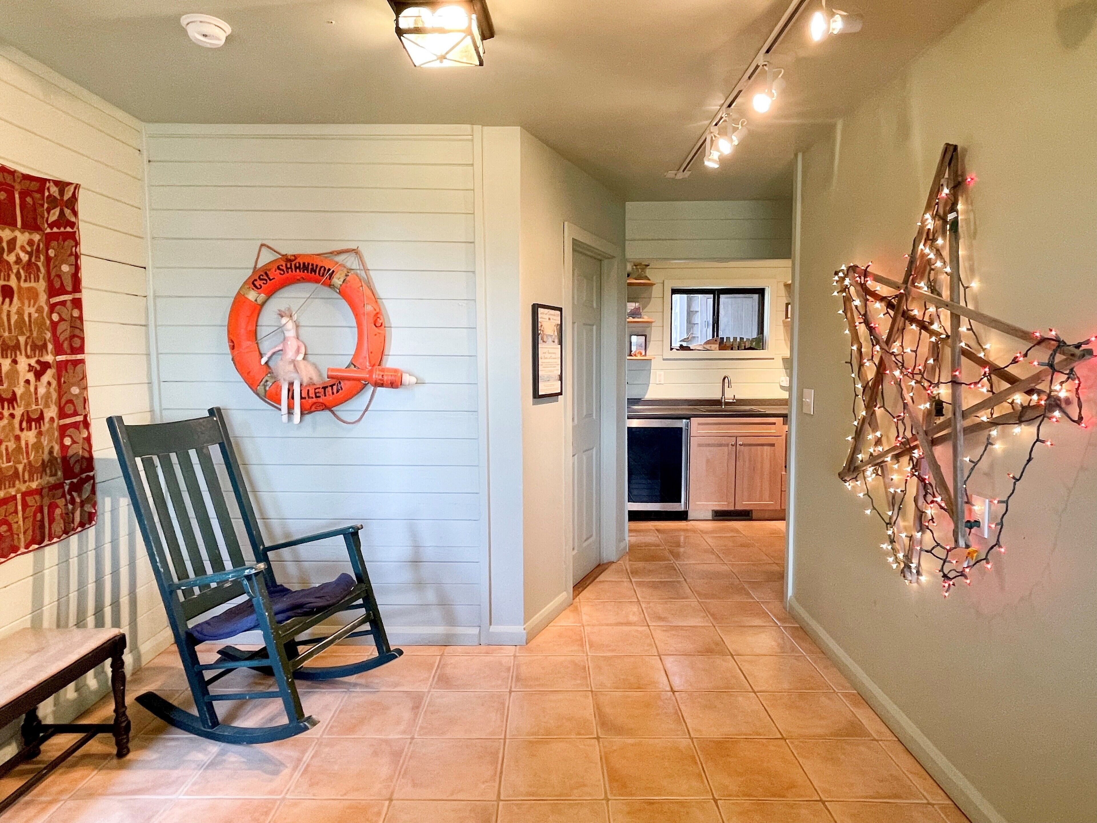 A hallway with a rocking chair, a life preserver decoration, and a kitchen visible in the background.