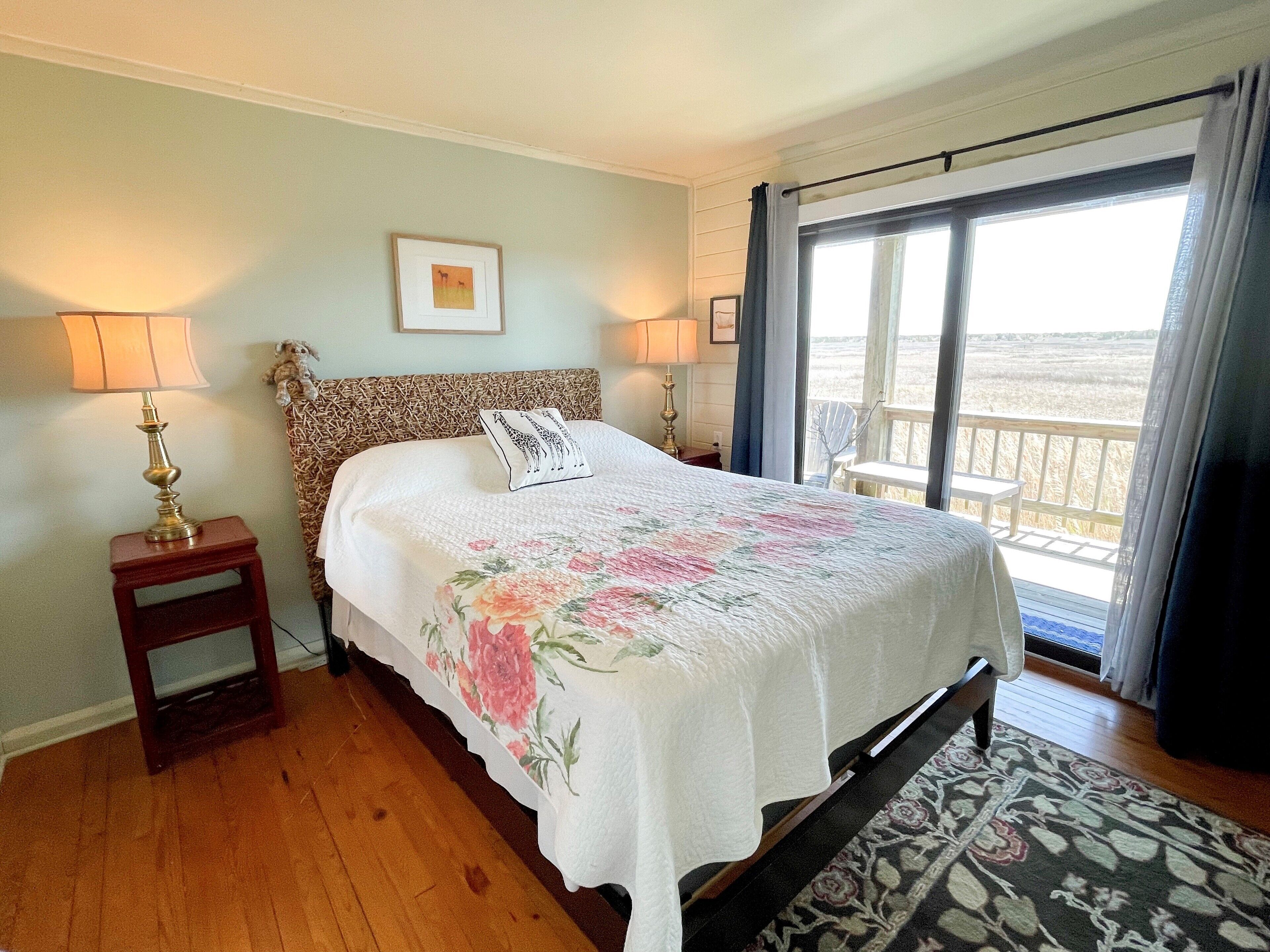 Een slaapkamer met een bed, twee bedlampjes, een ingelijst beeld aan de muur en uitzicht op het omliggende landschap door een raam.