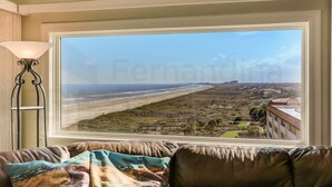 Blick aus einem Fenster auf einen Strand und ein Gebäude, im Vordergrund eine Lampe und ein Sofa.