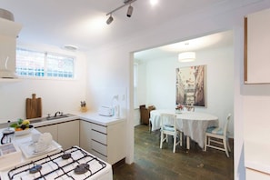 Cocina compacta con estufa a gas, lavamanos y ventana. Zona de comedor con mesa y sillas.