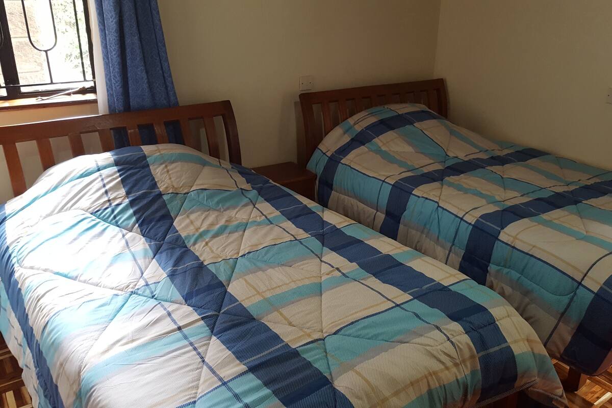 A room with two single beds, wooden headboards, a window with blue curtains, and a wooden floor.