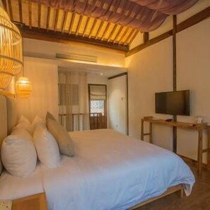 Un dormitorio con cama, almohadas, un televisor sobre una mesa de madera y una ventana con persianas.