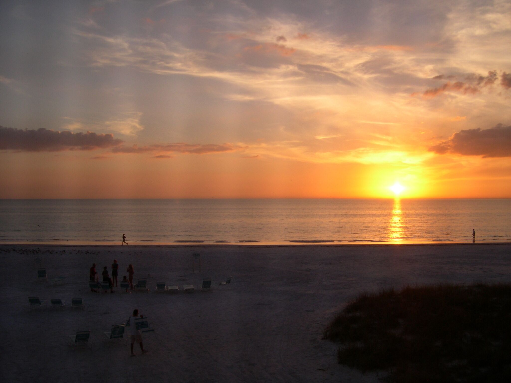 Een strand met mensen die wandelen en op stoelen zitten, en een zonsondergang boven de oceaan.