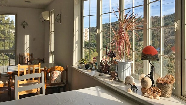 A cozy room with a large window, a dining table, chairs, and various decorative items on the windowsill. - Pension Smile Again (Shimoda)