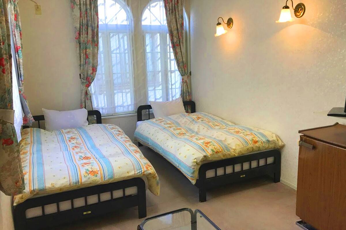 A room with two single beds, floral curtains, a wooden cabinet, and wall-mounted lights.