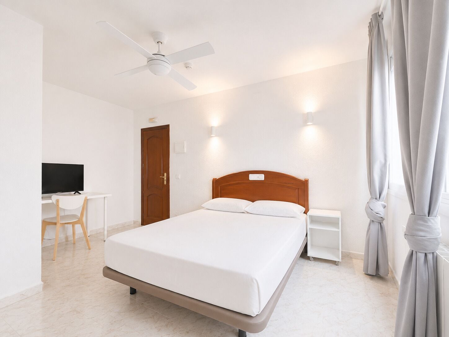 Une chambre d’hôtel comprenant un lit, un bureau avec une chaise, une télévision, un ventilateur de plafond et une fenêtre avec des rideaux.