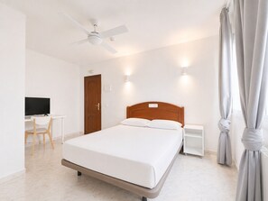 Habitación de hotel con cama, escritorio con silla, televisor, ventilador de techo y ventana con cortinas.