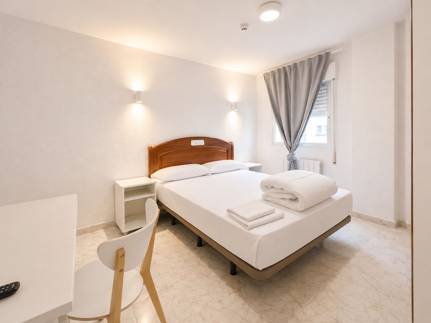 Una habitación de hotel con cama, mesita de noche, silla y ventana con cortinas.