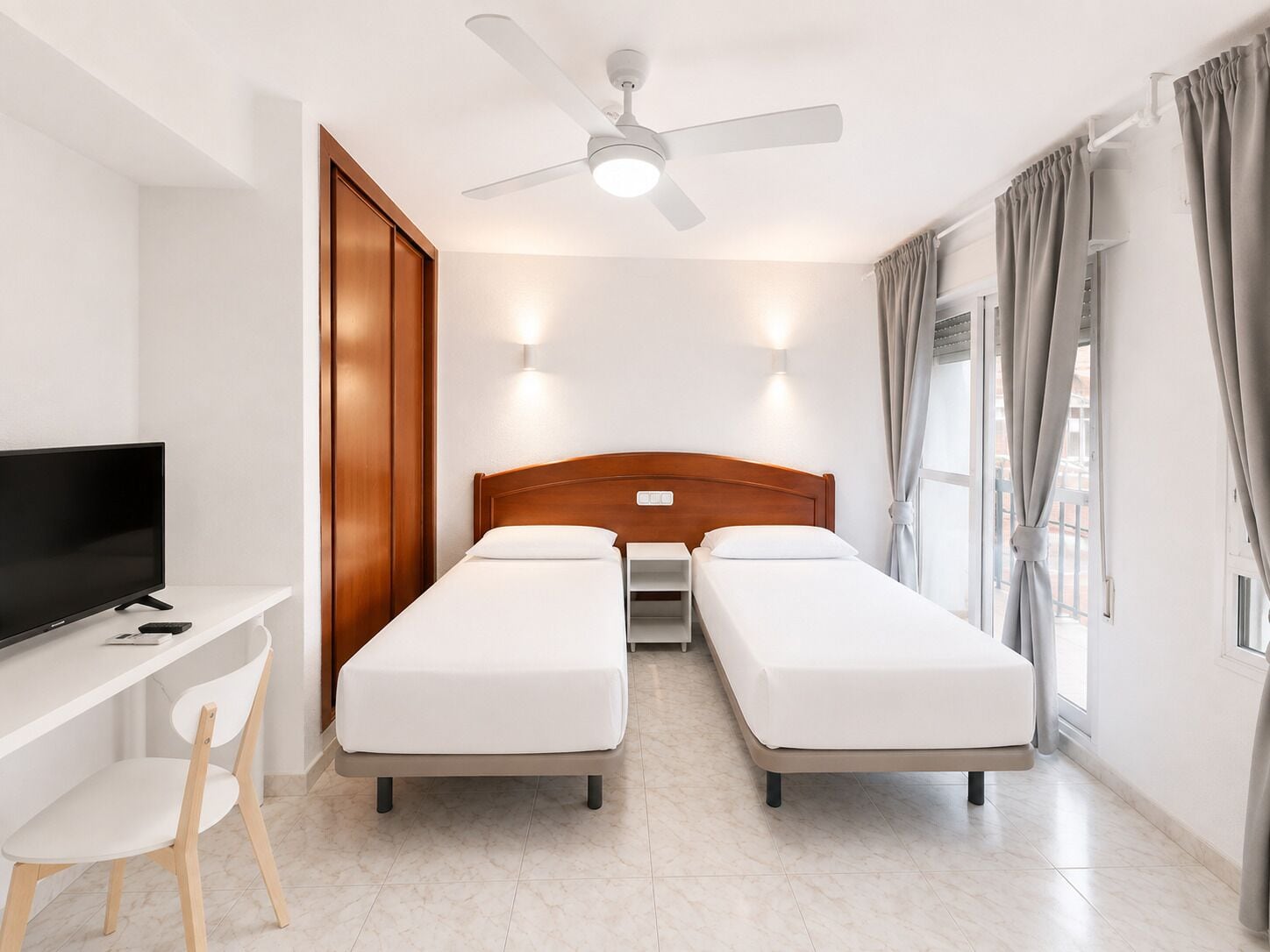 Una habitación de hotel con una cama, un escritorio con televisor, una silla, un ventilador de techo y una ventana con cortinas.