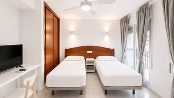 Una habitación de hotel con una cama, un escritorio con televisor, una silla, un ventilador de techo y una ventana con cortinas.