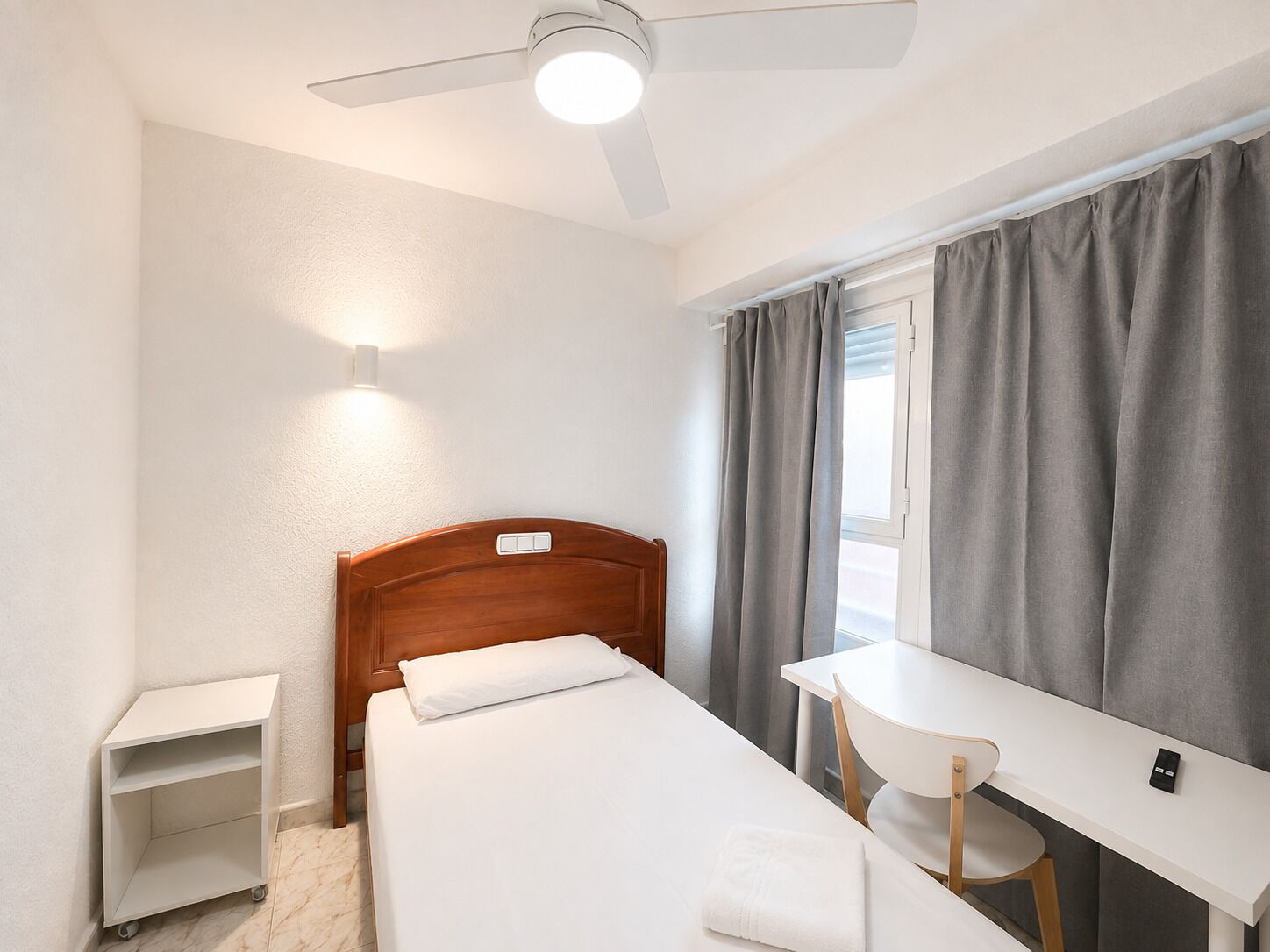A single bed with a wooden headboard, a bedside table, a chair, a desk, and a ceiling fan.