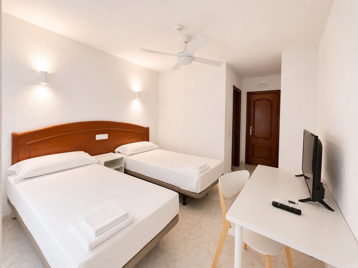 A hotel room with two beds, a desk, a chair, a television, and a ceiling fan.