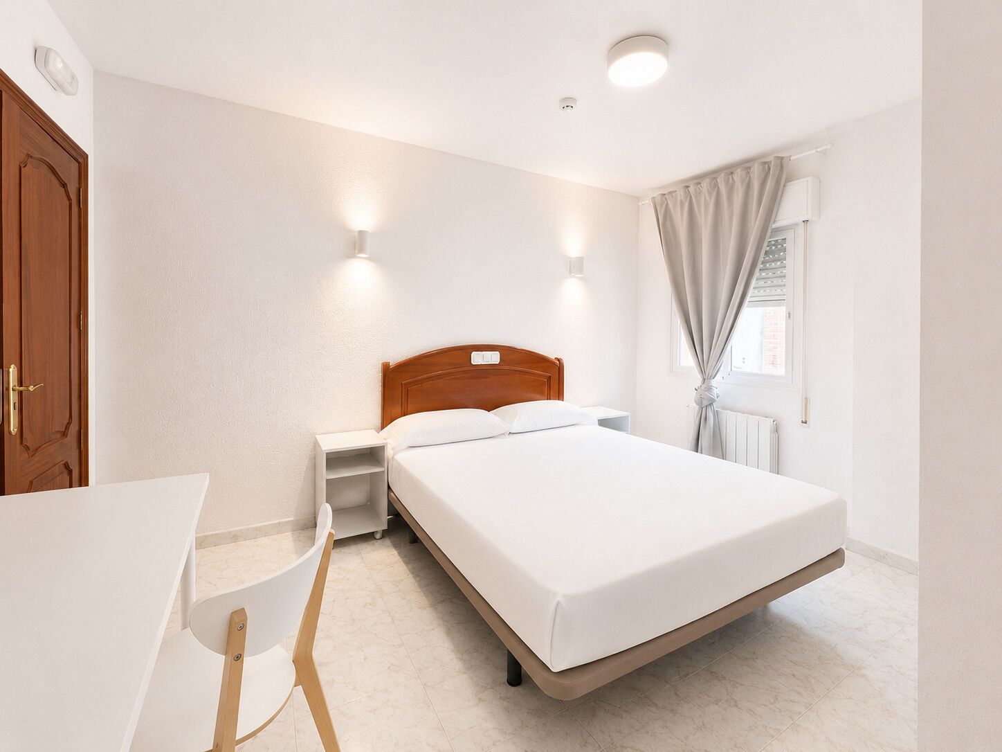 Een hotelkamer met een bed, nachtkastje, stoel en een raam met gordijnen.