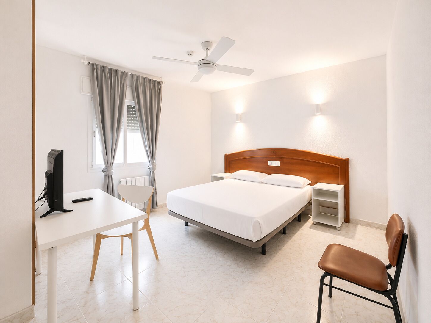 Habitación de hotel con cama, escritorio, silla, computadora, ventilador de techo y luces fijadas a la pared.
