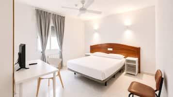 Habitación de hotel con cama, escritorio, silla, computadora, ventilador de techo y luces fijadas a la pared.